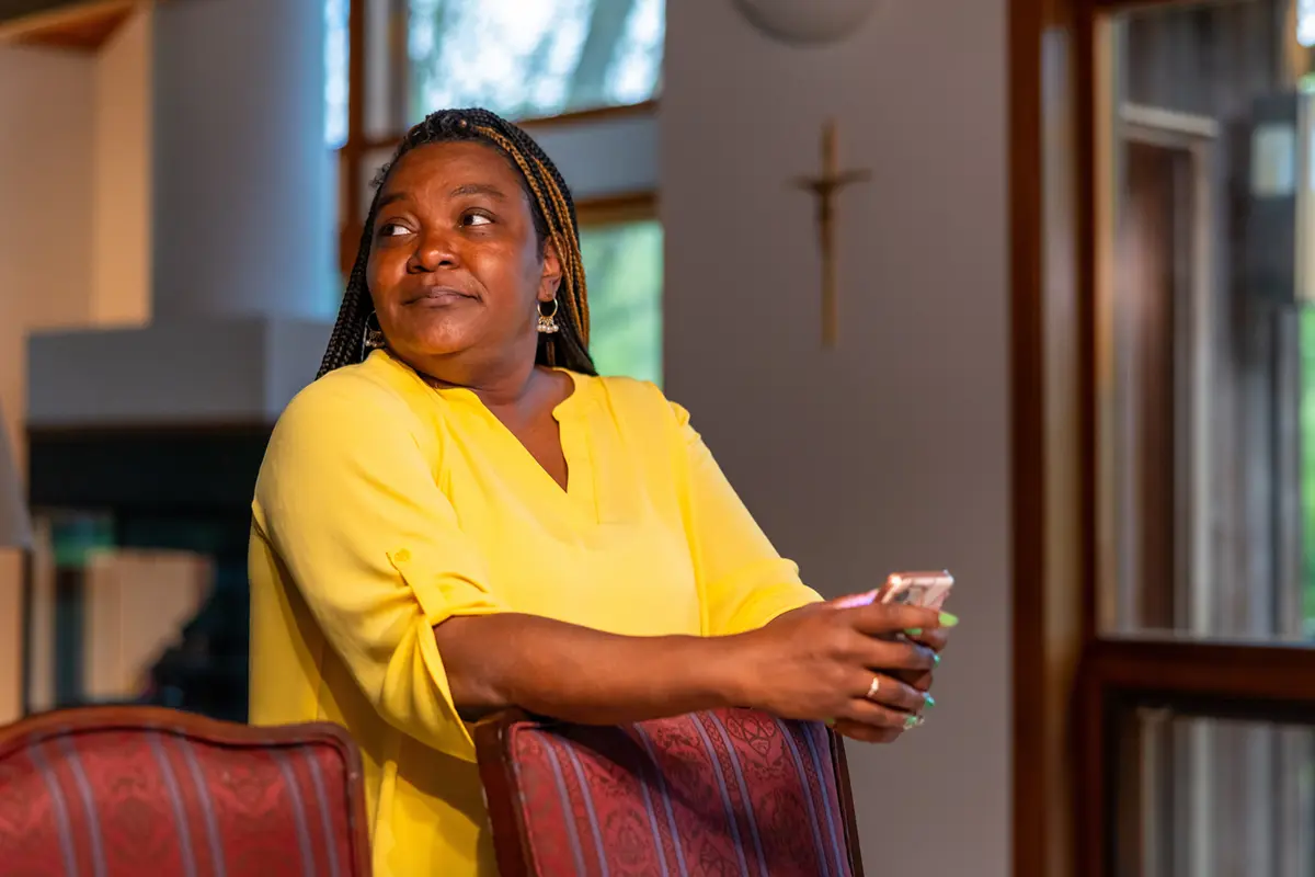 A dark-skinned woman in a yellow top leans on the back of a red upholstered chair while looking up and away.