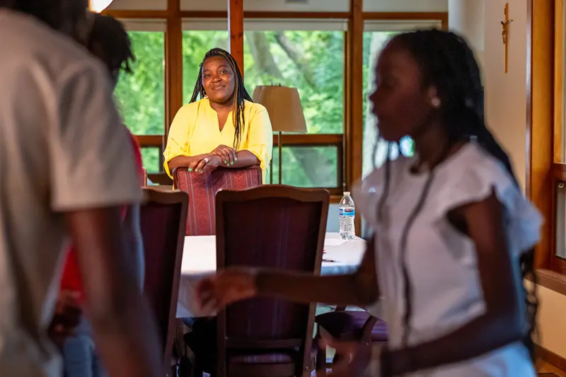 A woman in a yellow top stands behind a dining chair in a room with large windows overlooking green trees, watching two children.