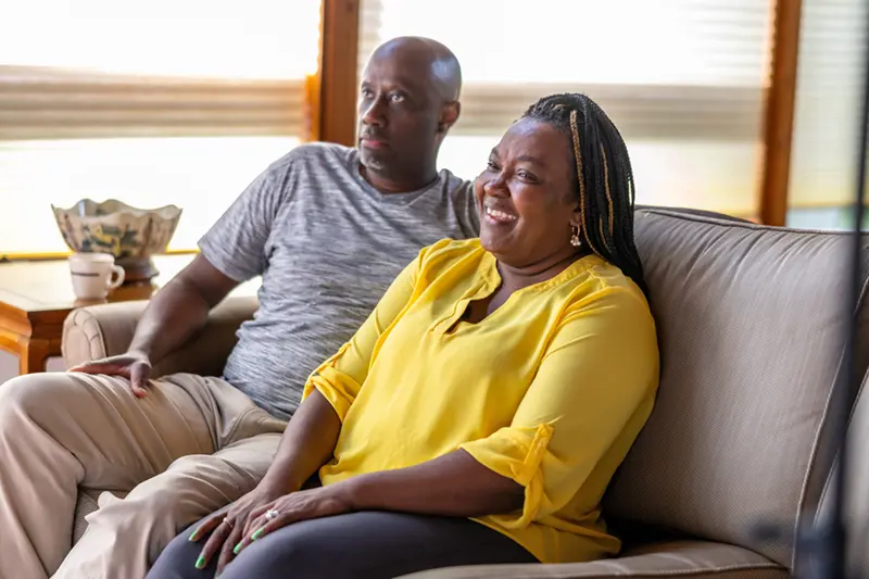 A dark-skinned woman with braided hair smiles while seated next to a man on a tan couch in a home living room.