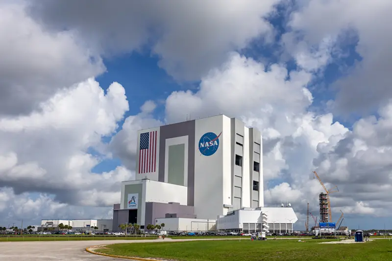 The Vehicle Assembly Building at the NASA Kennedy Space Center under a partly cloudy blue sky.