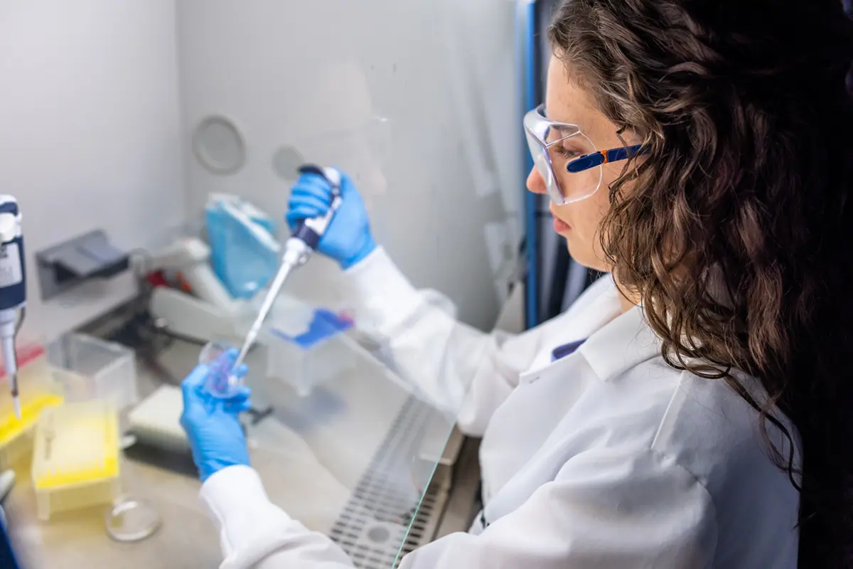 A young woman with dark, curly hair in a lab coat and safety goggles pipettes liquid in a laboratory hood.
