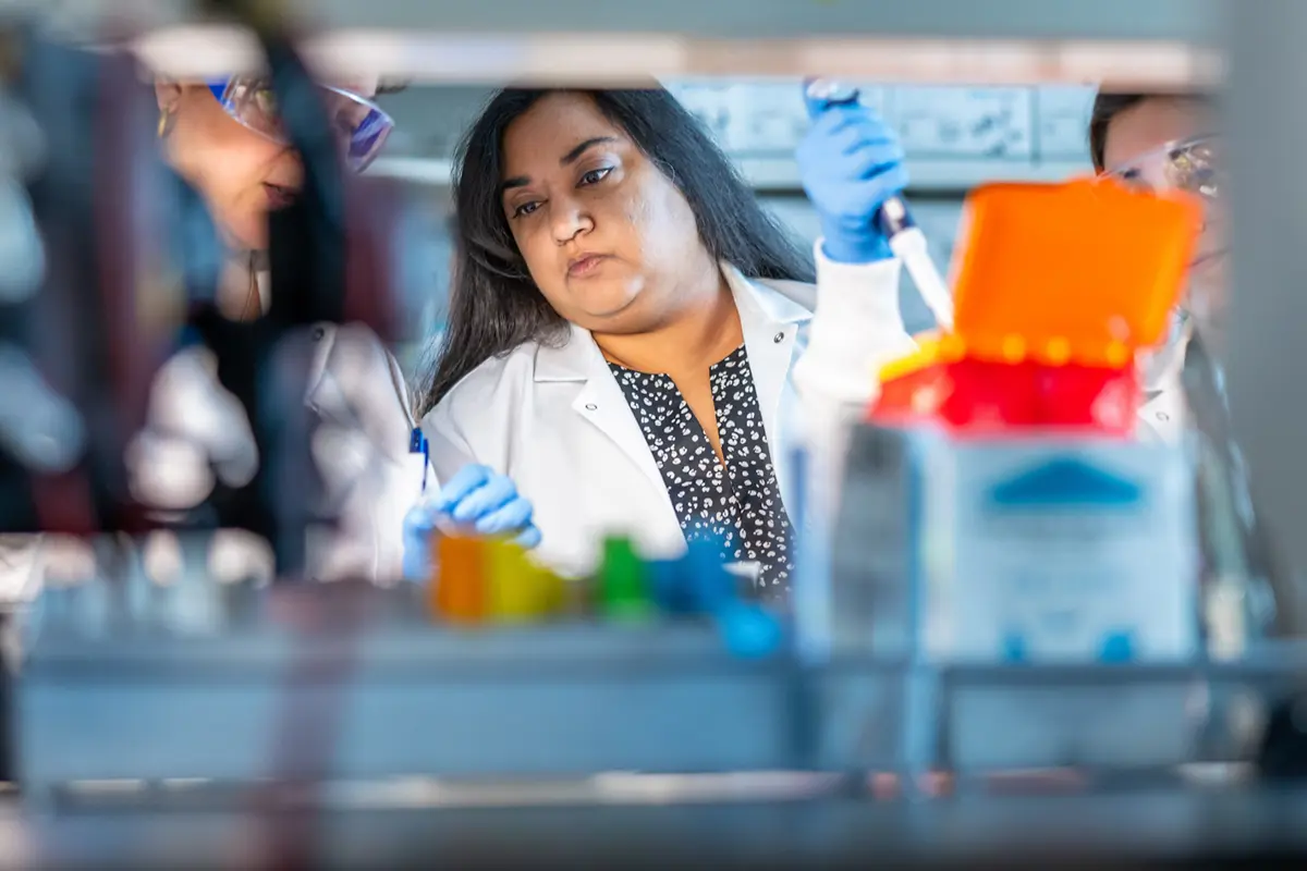 A dark-skinned woman in a white lab coat and blue gloves pipettes liquid while working with two other people in a laboratory.