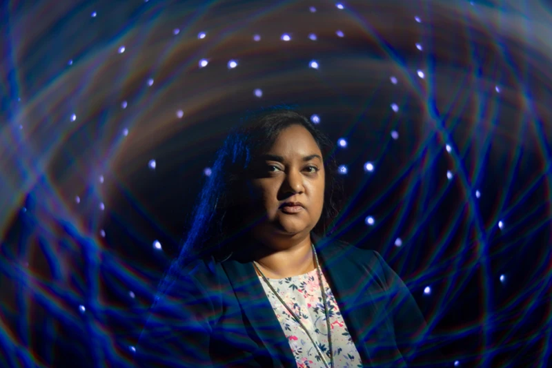 A dark-skinned woman with dark hair wears a floral shirt and a black blazer, surrounded by swirling blue lights.