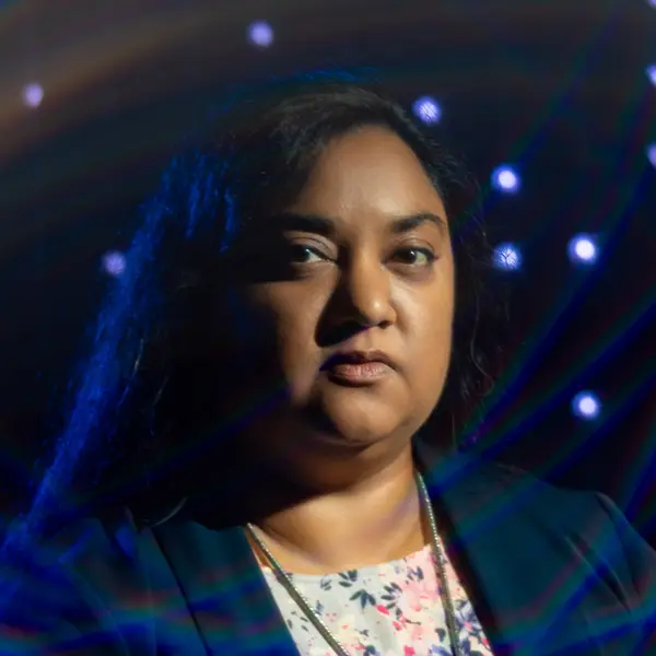 A headshot of a dark-skinned woman with dark hair, wearing a blazer, surrounded by blurred blue and white lights.