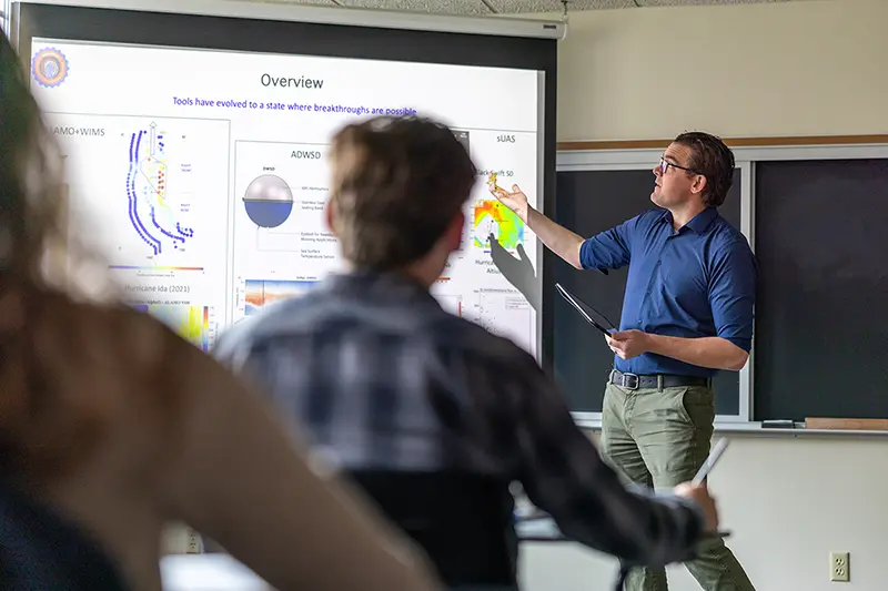 David Richter standing in front of a classroom referencing a projector screen while teaching class.