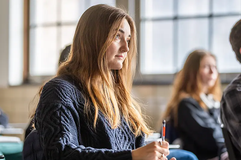 A female student looks up while taking notes, listening to David Richter teach class.