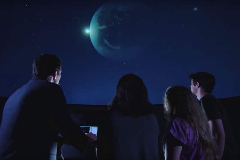 A profressor and three students look at planets on a large screen.