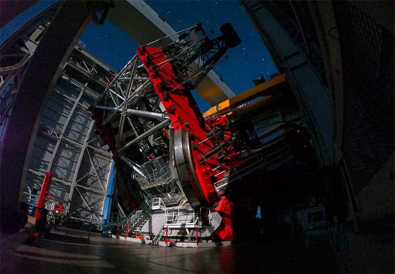A lage binocular telescope sits inside of an observatory. Stars glow in the night sky.