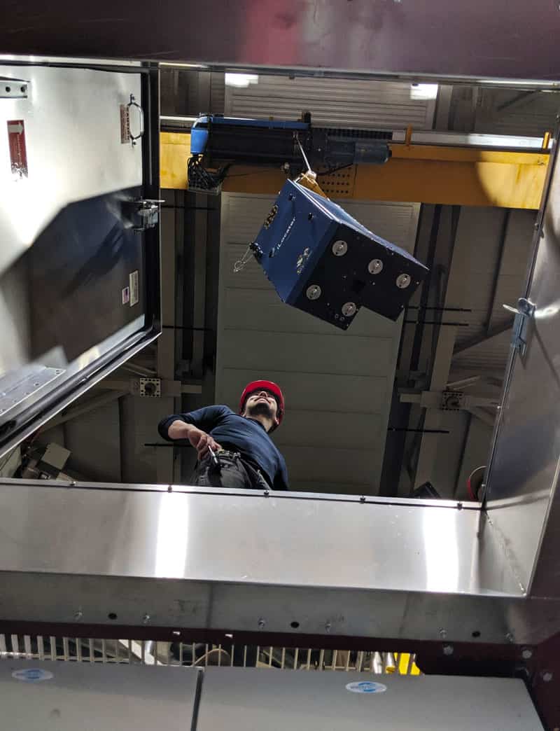 A man weaing a hard hat watches the iLocater, inside a blue paneled box, lower into an area where it will be installed.