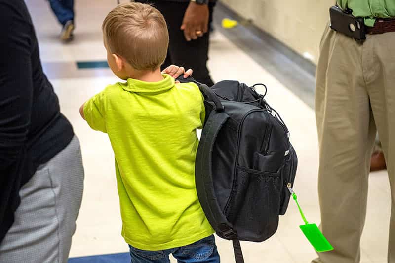 A child walks down a hallway holding a backpack on his shoulder.