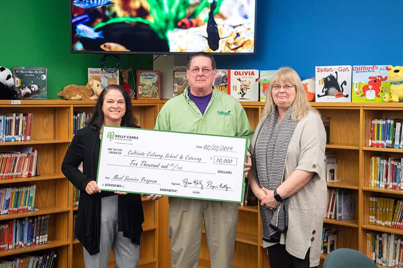 Three people stand in a classroom, in front of a wall of childrens books, holding a check for 10,000 dollars.