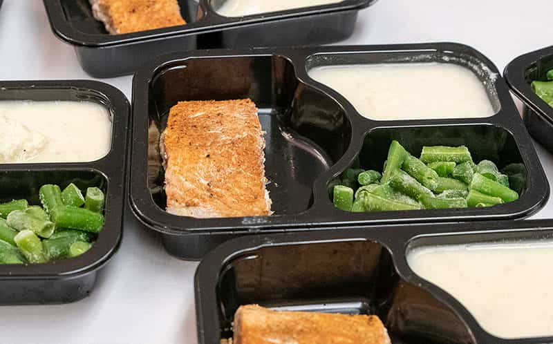 Salmon, green beans, and mashed potatoes divided in black plates and ready to be packaged.