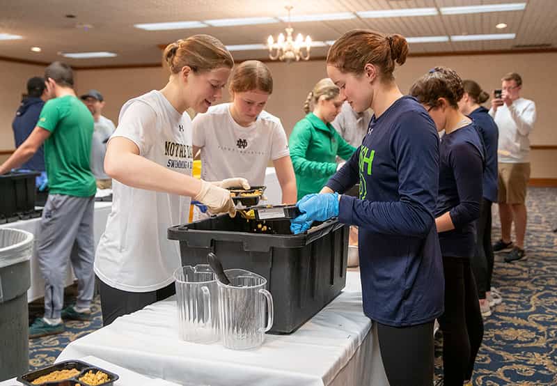 Several student athletes, wearing hair nets and gloves, scoop food in divided plates, preparing them to be packaged.