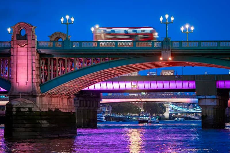 A image of a blurry red double-decker buse as it travels on a bridge over the River Thames at night.