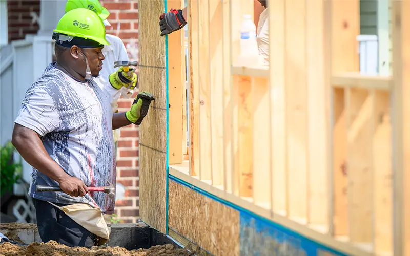 A construction worker wearing a lime green Habitat for Humanity hardhat uses a hammer to build the frame of a house.	He's wearing work gloves and holding the hammer in his left hand.