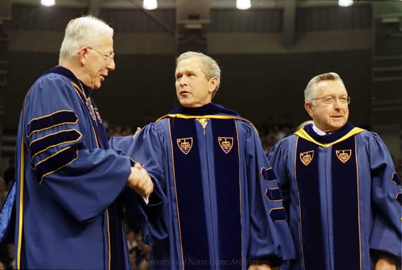 Bush and two men dressed in graduation regalia shake hands at commencement.