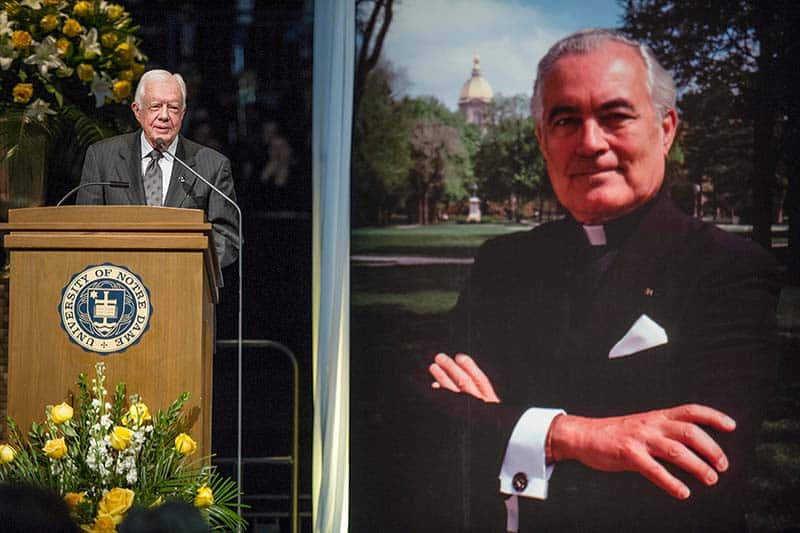Jimmy Carter speaks at a podium beside a large poster of Father Hesburgh.