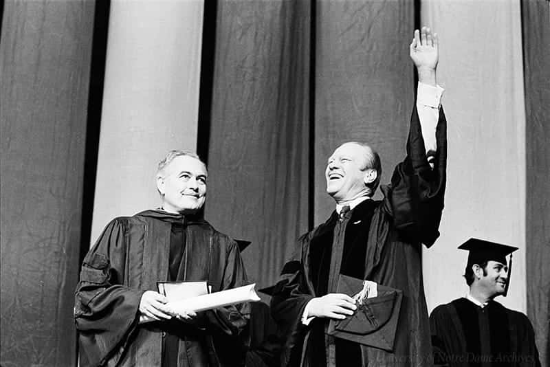 Father Hesburgh stands smiling next to Gerald Ford as he waves smiling.