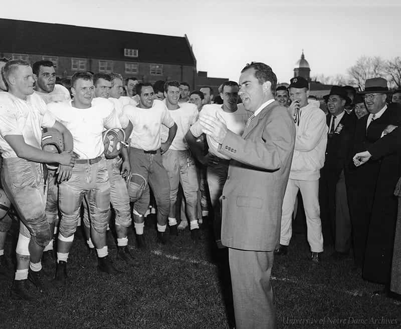 Richard Nixon speaks to a group of Notre Dame football players, all smiling. The Golden Dome is visible in the background.