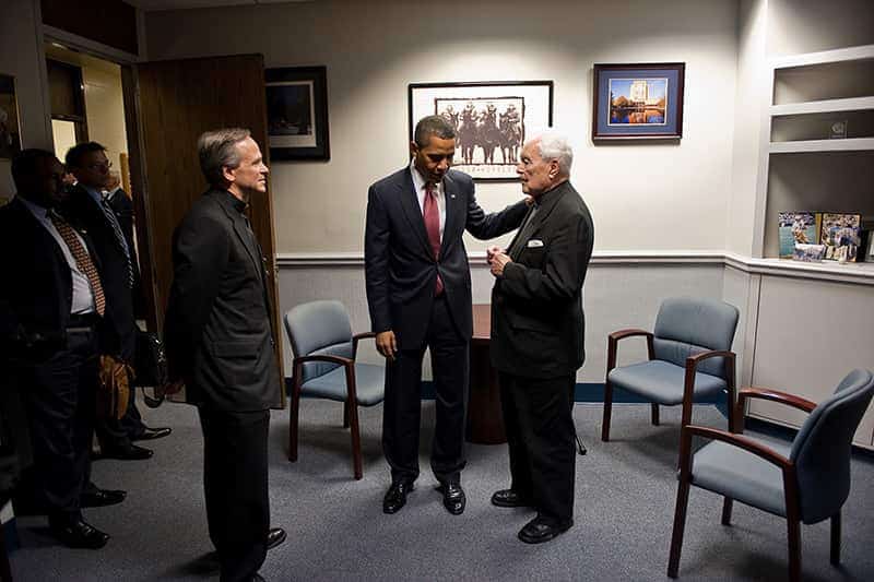 Barack Obama puts his hand on Rev. Theodore M. Hesburgh's shoulder while they have a discussion.	Rev. John Jenkins, C.S.C. stands beside them.