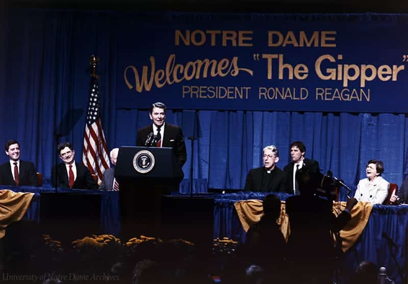 Ronald Regan stands at a podium on stage in front of a sign that reads Notre Dame Welcomes The Gipper President Ronald Reagan.