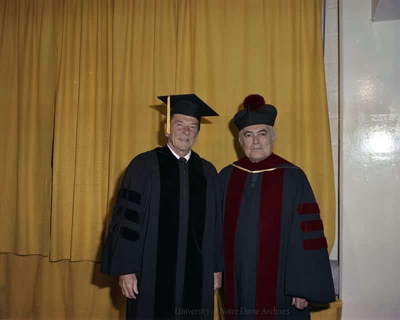 Ronald Reagan and Father Hesburgh smile for the camera wearing graduation regalia in front of gold curtains.