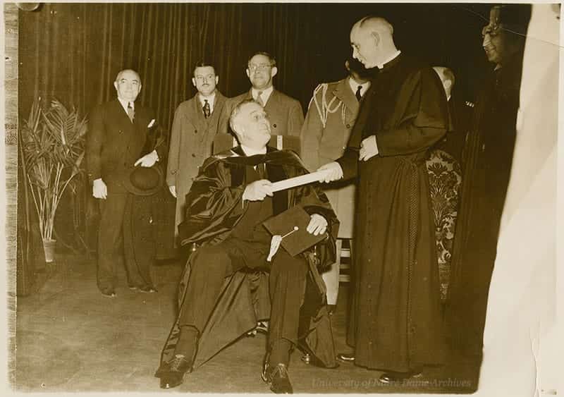 Roosevelt sits in a chair and accepts an honorary degree from a priest.