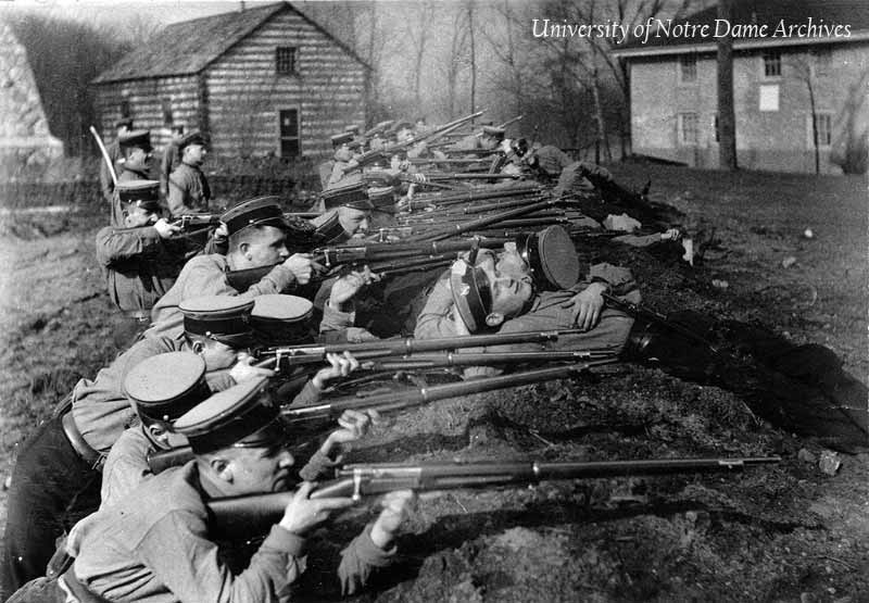 Students in the military training corps at rifle target practice in the trenches, 1915-1916.	The Founder's Monument, Log Chapel, and Old College are in the background.