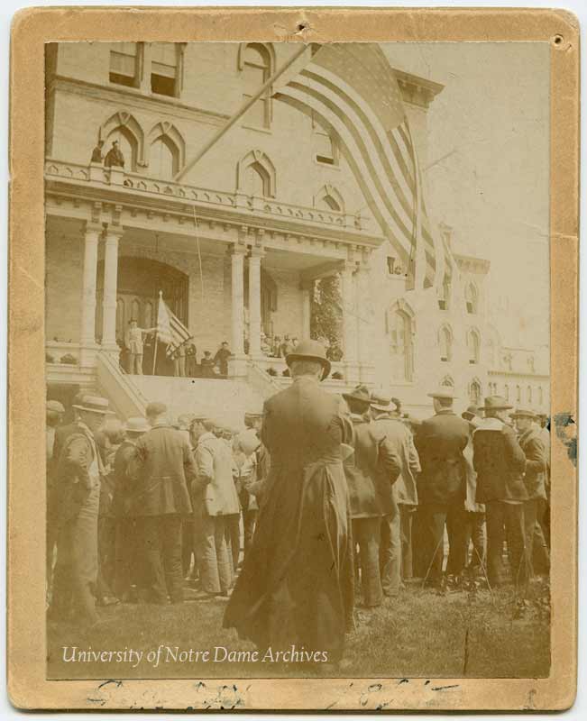 Decoration Day - Colonel William Hoynes addressing the student body from the porch of Main Building with an American flag in hand and flying from the building, 1896.