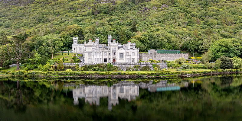 View from across the lake of Kylemore Abbey, a large light gray colored stone building with a tall tower, surrounded by trees and hills.