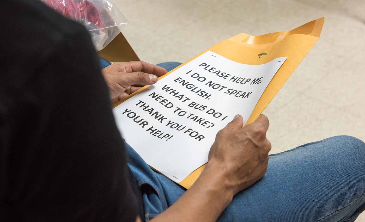 A man sitting on a chair with a sign reading &ldquo;Please help me. I do not speak English. What bus do I need to take? Thank you for your help!&rdquo;