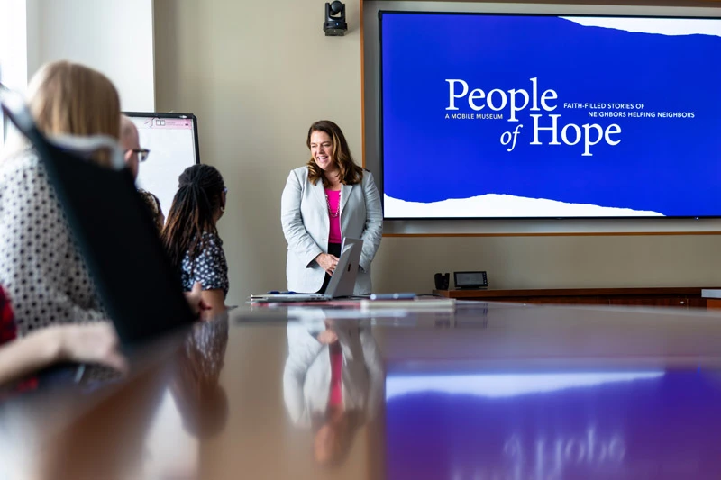A woman in a light gray blazer and pink shirt smiles at a small group seated around a conference table.  Behind her, a large screen displays the 'People of Hope' mobile museum logo on a blue background.