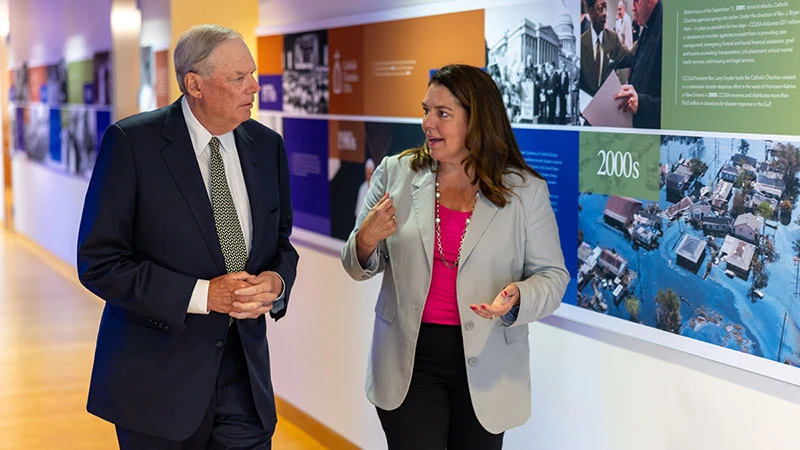 A man in a dark suit and tie and a woman in a light gray blazer and pink shirt walk and talk in a hallway with a historical photo display on the wall.  The display includes a photo of Hurricane Katrina flooding.
