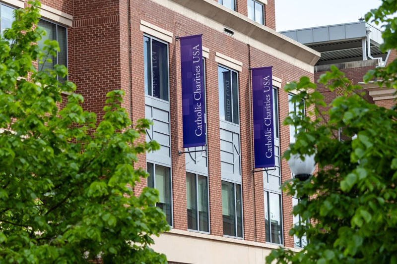 Purple Catholic Charities USA banners hang on a brick building, partially obscured by green trees.