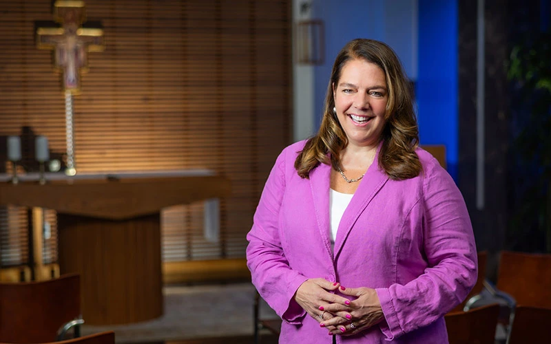 A woman with shoulder-length brown hair, wearing a fuchsia blazer and white shirt, smiles with her hands clasped in front of her in a small chapel. A crucifix and altar are visible in the background.