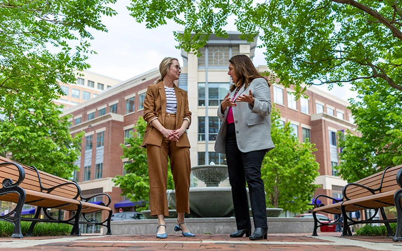 Two women converse near a fountain on the brick walkway outside of Eddy Street Commons.  One woman wears a brown pantsuit and the other a light gray blazer and black pants. Lush green trees frame the red brick buildings behind them.