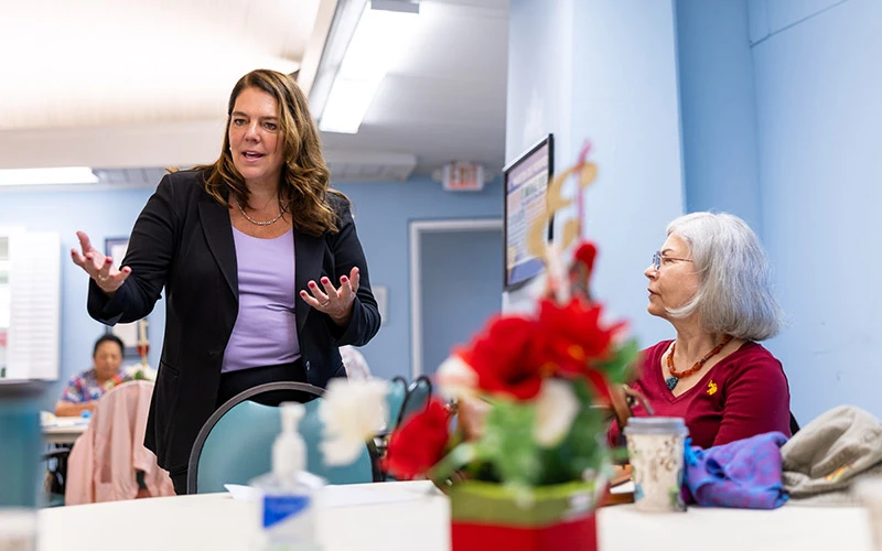 A woman in a black blazer and purple shirt gestures with her hands as she speaks to a woman with short gray hair wearing a maroon shirt.  A poinsettia in a red pot sits on the table between them.