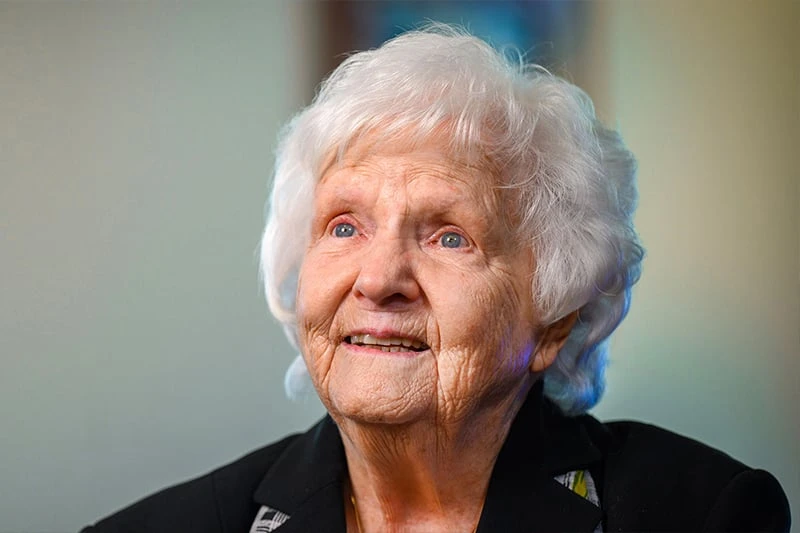 An elderly woman with short, white hair and bright blue eyes looks upward with a slight smile. She wears a dark jacket.