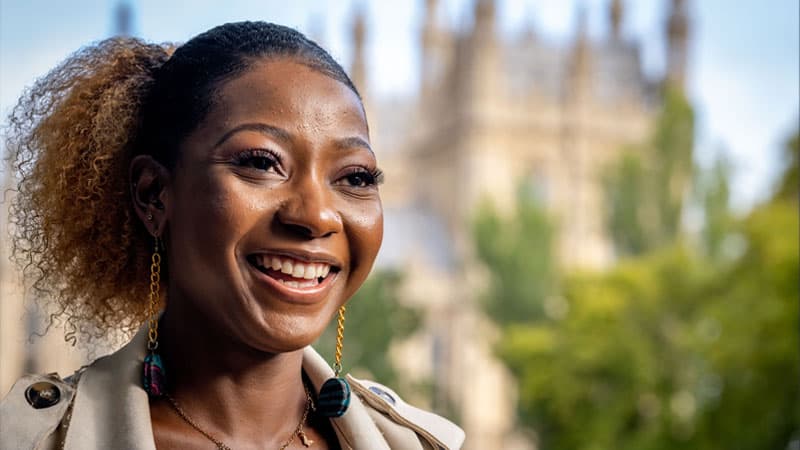 A portrait of Bupe Lughano Kabaghe
 with a blurry view of the Parliament building in London in the background.