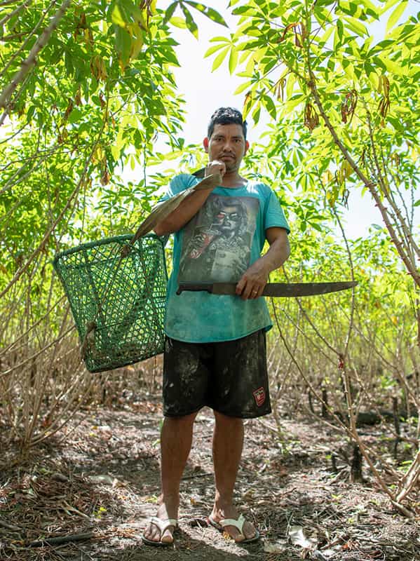 A man holds a basket of farinha roots in one hand and a machete in the other.