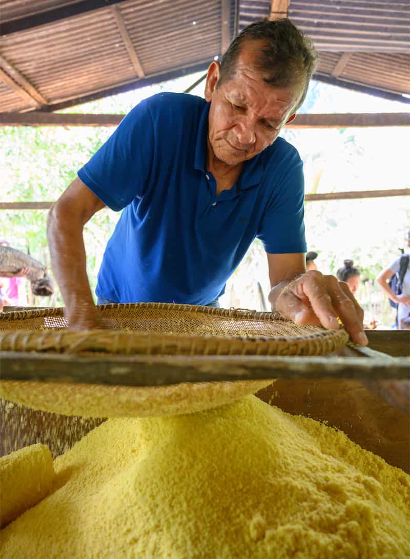 A man pushes dried flour clumps through a woven sieve.