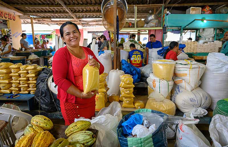 A smiling woman at a market holding a clear bag of farinha surrounded by other farinha bags and fruits.