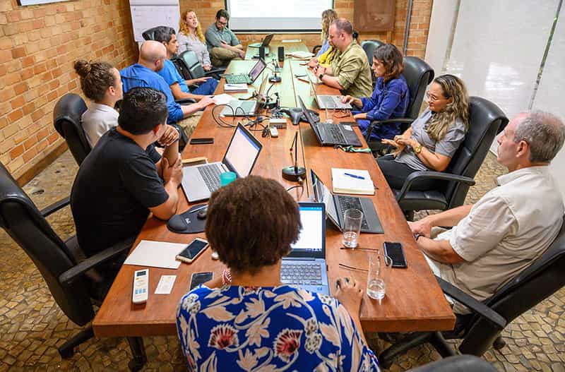 People in a conference room sit around a table talking. Laptops, notepads, and other miscellaneous items are on the table.