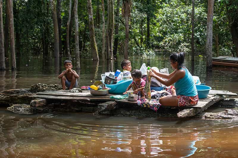 A mother washes dishes and clothing in a flooded area as her three children sit nearby.