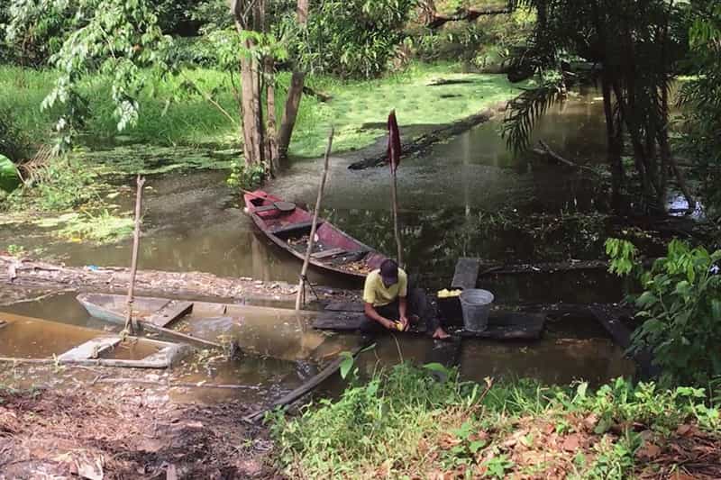 Placeholder image for a video - a man sits on a dock and washes fruit in water, a boat floats behind him.