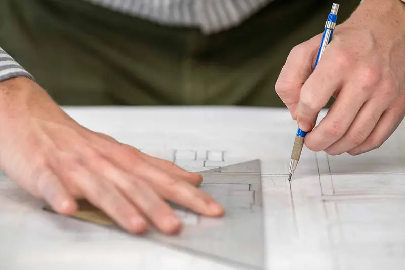 A close up of someone creating a architectual sketch. One hand holds onto a triangular ruler and the other draws a straight line with a pencil.