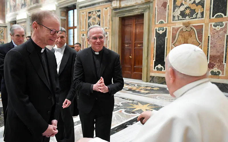 Two priests in black suits and clerical collars clasp their hands and smile as they speak with Pope Francis, who is seen from behind wearing his white zucchetto and cassock.