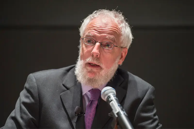 A man with white hair and beard, wearing a dark suit and purple patterned tie, speaks into a microphone.