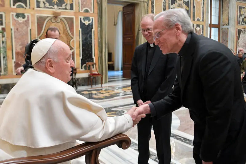 Pope Francis shakes hands with a man in a black suit and clerical collar, while another priest looks on.