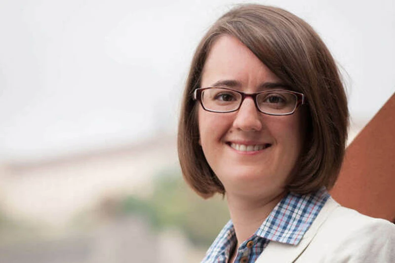 Woman with shoulder-length brown hair, glasses, and a blue and red plaid shirt smiles.